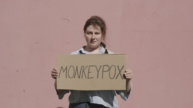 Sad, tired young woman stands near pink wall, turns and shows cardboard poster with covid-19 and monkeypox inscriptions. Sunny day, the wind blows the hair of a plus size lady.