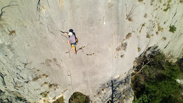 AERIAL: Young male rock climber lead climbing and using quickdraw to attach rope. Male climber in the middle of sunlit limestone wall hooking the rope. Outdoor sport activity in natural environment.