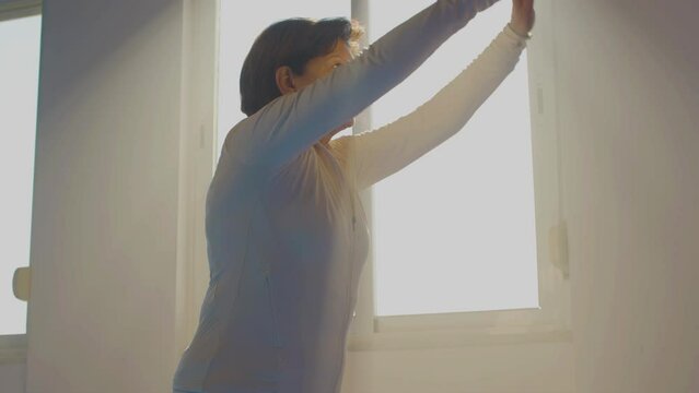 Senior woman practicing tai chi in gym. Medium shot of focused woman in sportswear performing slow movements in sunlight, doing physical activity indoor. Chinese martial arts, sport, fitness concept