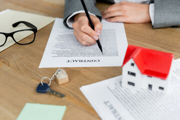 cropped view of woman signing contract near key chain with key and house model on desk.