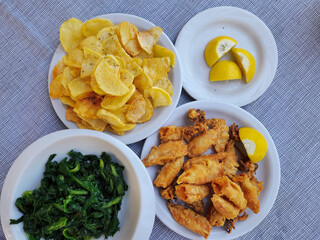 Top view of served tavern food table. Close up on dishes with fried calamari portion with slices of lemon, fries and leafy vegetables on restaurant in Greece.