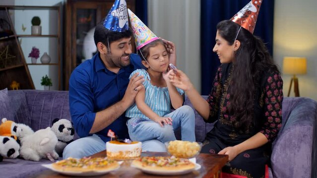 An Indian couple wearing a colorful hat celebrating their daughter's birthday -birthday party. Indian husband-wife feeding a cake to their adorable girl child - cake cutting  family bonding  loving...