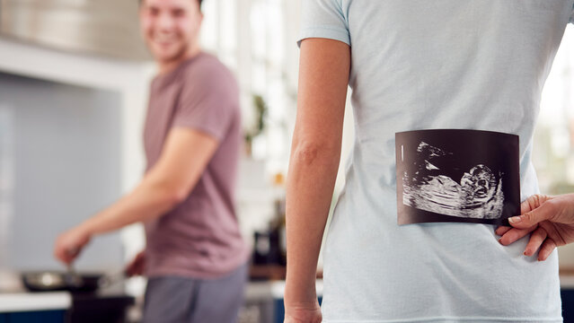 Pregnant Transgender Couple At Home In Kitchen With Woman Surprising Man With Ultrasound Scan Of Baby