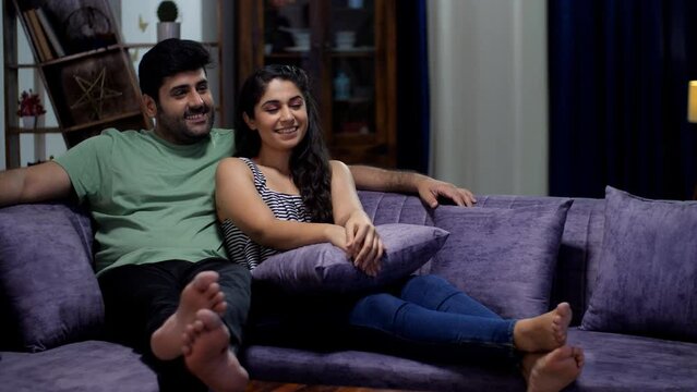 A young happy couple sitting on a comfortable sofa and watching television together - boyfriend and girlfriend  fun and entertainment. Newly married couple spending quality time watching OTT progra...
