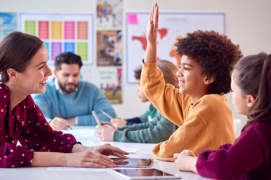 Group Of Multi-Cultural Students With Teachers In Classroom Putting Hand Up To Answer Question