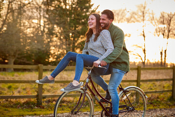 Fototapeta na wymiar Couple Riding Bike Through Countryside With Woman Sitting On Handlebars