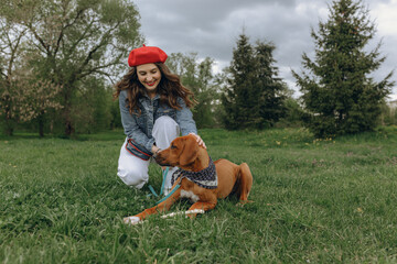 Positive stylish woman in red beret petting brown dog while sitting on grassy field in nature together 