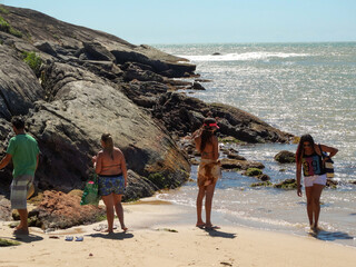 Linda praia, com pessoas passeando, grandes rochas, banhada pelas ondas, muita vegetação e...