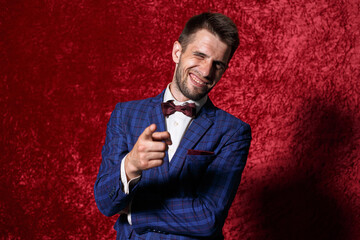Cheerful well dressed businessman in suit and bow tie pointing at camera with index finger and showing want gesture while winking and looking at camera on red background in studio 
