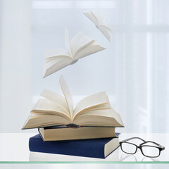 Time to read your favorite books. Old books and black glasses on a glass shelf against open window with transparent curtain and flying books