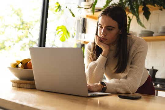 Thoughtful non-binary trans woman using laptop in the kitchen at home