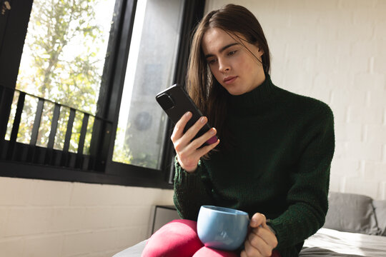 Non-binary trans woman holding coffee cup using smartphone in bedroom at home
