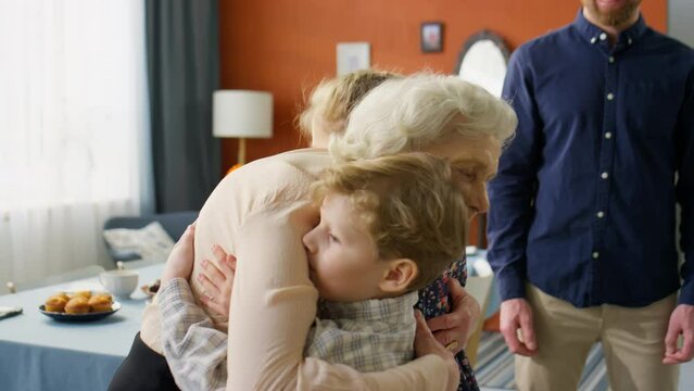 Joyous elderly grandmother giving hugs to little grandkids while greeting family at home for holiday dinner