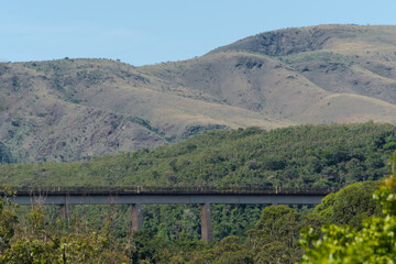 Obraz premium Vista de longe, linha ferrea construída sobre ponte aos pés de montanha, cercado de muita vegetação e lindo céu azul, na região do Parque das águas, localizado no Barreiro, Belo horizonte, Minas Gerai
