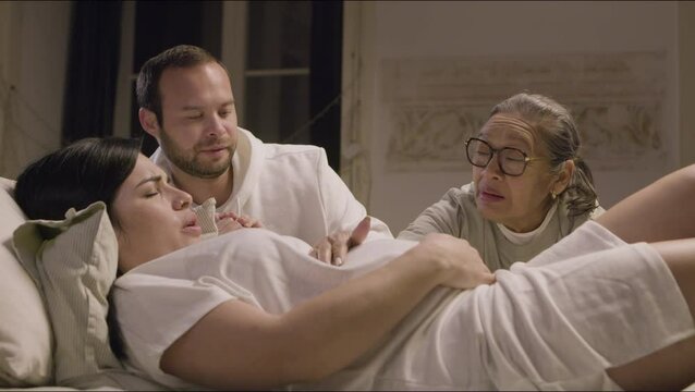 Pregnant woman lying on bed in bedroom and having contractions . Husband and Asian doula talking with expecting mother, comforting her and stroking belly. Parenthood, medicine, pregnancy concept