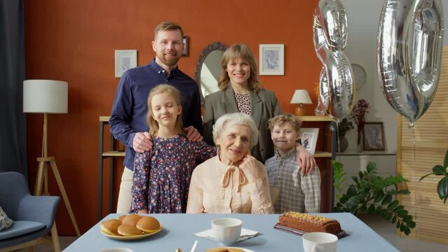 Zoom in family portrait of cheerful elderly grandmother sitting at dinner table and posing for camera with family while celebrating birthday
