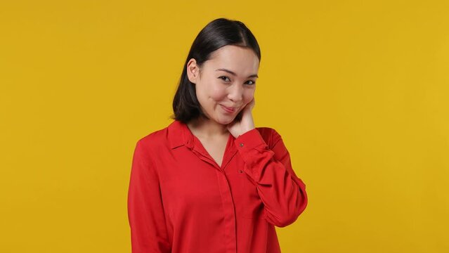 Smiling good kind surprised happy cheerful young woman of Asian ethnicity 20s years old wears red shirt ask who me oh it so sweet put hands on chest isolated on plain yellow background studio portrait