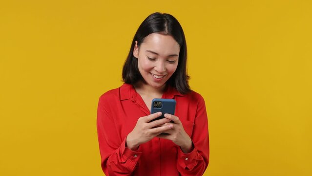 Happy excited fancy young woman of Asian ethnicity 20s wears red shirt hold in hand use on mobile cell phone chatting texting sending sms gesture isolated on plain yellow background studio portrait