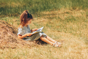 Serious child girl in a dress reads a book on a haystack, schoolgirl rural life, sunny summer day