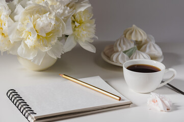 Open white notepad, cup of coffee, sweets and a bouquet of peonies on a white table. Freelancer or blogger workplace concept.