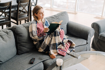 Young white woman wrapped in blanket reading book sitting on sofa