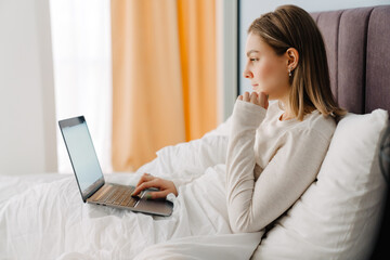 Young white woman using laptop while lying in bed at home in morning