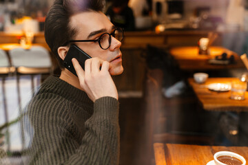 Young man smiling while drinking coffee and using cellphone in cafe