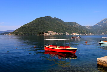 Fototapeta na wymiar Boka Kotor, Montenegro. A red boat sails along Bay Kotor against backdrop of famous island and Church Madonna on Reef, Perast