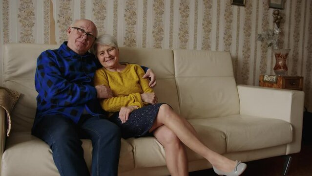 elderly caicasion couple looking at the camera together while higging and sitting on the sofa. High quality 4k footage