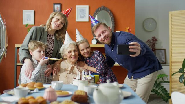 Cheerful family members in party hats taking selfie with happy senior grandmother on her 80 years old birthday celebration at home