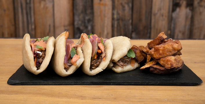Asian cuisine. Closeup view of pulled pork baos buns with pickles, sauce and fried sweet potatoes, in a black dish on the wooden table