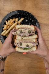 Top view of caucasian female hands holding a a Vitel tonné sandwich with veal, cream, capers, arugula and french fries. - obrazy, fototapety, plakaty
