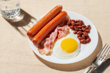 Eggs with bacon, sausages and beans in a plate on the table. Hard sunlight