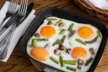 Fried eggs with vegetables (mushrooms and asparagus) in a small frying pan with cutlery, napkins and a wicker basket on wooden background. Breakfast idea or concept, close up