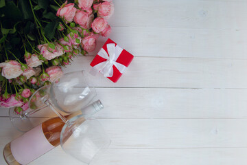 A large bouquet of pink roses, a gift in a red box and a bottle of rose wine with glasses lie on the side on a wooden white background with a place for text. High quality photo - Powered by Adobe