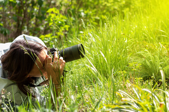Professional female photographer taking a photo in nature while lying down on the green grass.
