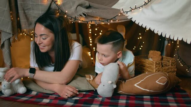 Mom and little son play toys while lying in a tent of blankets and pillows in the bedroom. A family