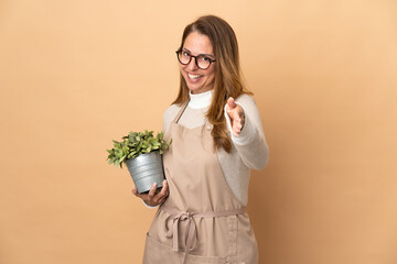 Middle age gardener woman holding a plant isolated on beige background shaking hands for closing a good deal