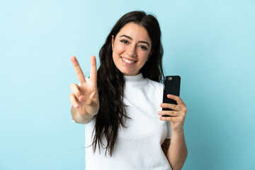 Young woman using mobile phone isolated on blue background smiling and showing victory sign