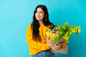 Young woman holding a grocery shopping bag isolated on blue background having doubts and thinking