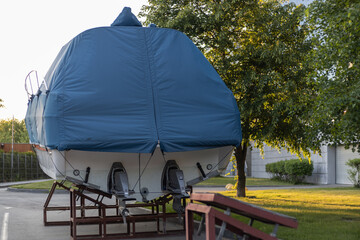 A motor boat covered with a blue cover stands on land under the open sky against the background of trees