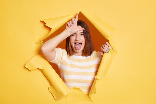 Indoor shot of excited attractive woman stands in torn paper hole, showing looser gesture with hand on forehead, looking through breakthrough of yellow background.