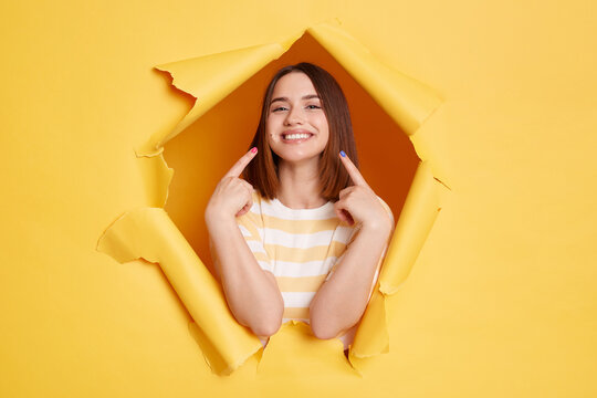 Indoor shot of smiling young adult woman stands in torn paper hole, pointing at her perfect smile and teeth, looking through breakthrough of yellow background.
