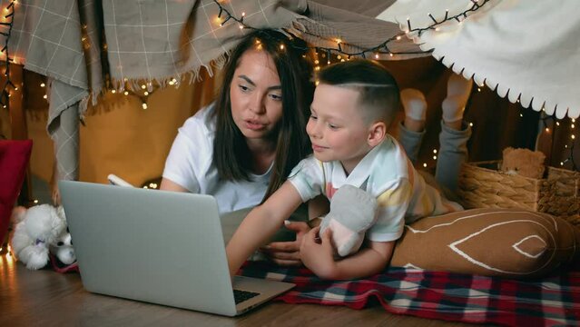 Mom and little son are watching a cartoon on a laptop while lying in a tent of blankets and pillows in the bedroom. A family