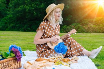 Girl in polka dot dress and hat sitting on white knit picnic blanket plays ukulele and drinking...