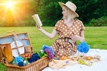 Girl in polka dot dress and hat sitting on white knit picnic blanket reading book and drinking...