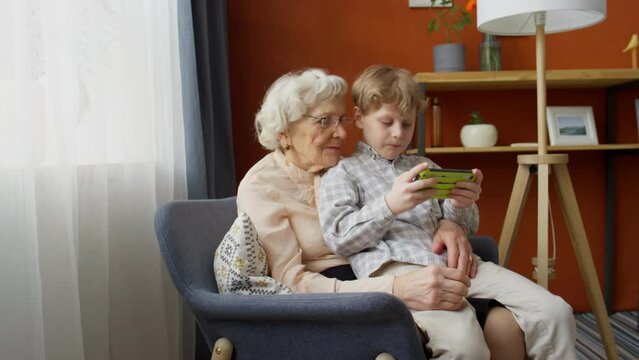 Zoom in shot of little boy sitting on knees on grandmother at home and playing video game on portable console