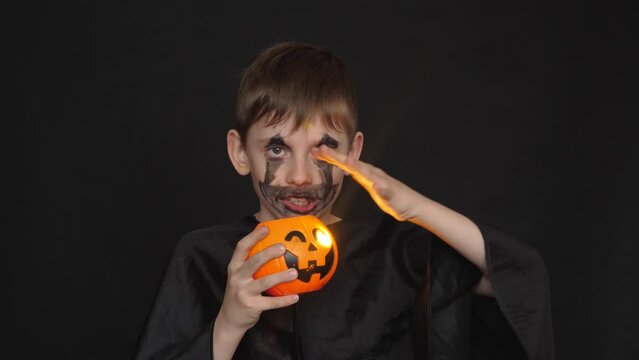 a mystical scary image of a cute caucasian boy 7-8 years old in a black costume for the Halloween holiday. a child painted in a terrible grime with a pumpkin. studio shooting, black background