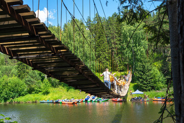 People in life jackets cross the suspension bridge over the Chusovaya River. Parking of colorful boats and catamarans for rafting. Active lifestyle and sports - obrazy, fototapety, plakaty