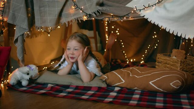 A little girl looks at the camera while lying in a tent of blankets and pillows in the bedroom. Children